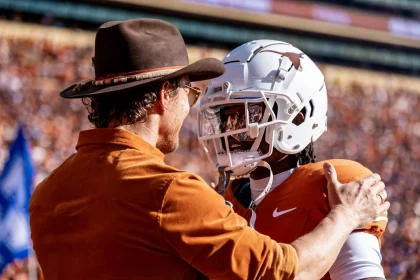 Isaiah Bond - Ein Mann mit braunem Hut und Hemd umarmt Isaiah Bond herzlich. Er trägt eine orange-weiße Uniform und einen Helm mit Longhorn-Logo. Sie stehen auf dem Footballfeld, umgeben von verschwommenen Zuschauern, die die lebhafte Spieltagsatmosphäre einfangen. Diese Beschreibung wurde mit der FootballR KI automatisch generiert.