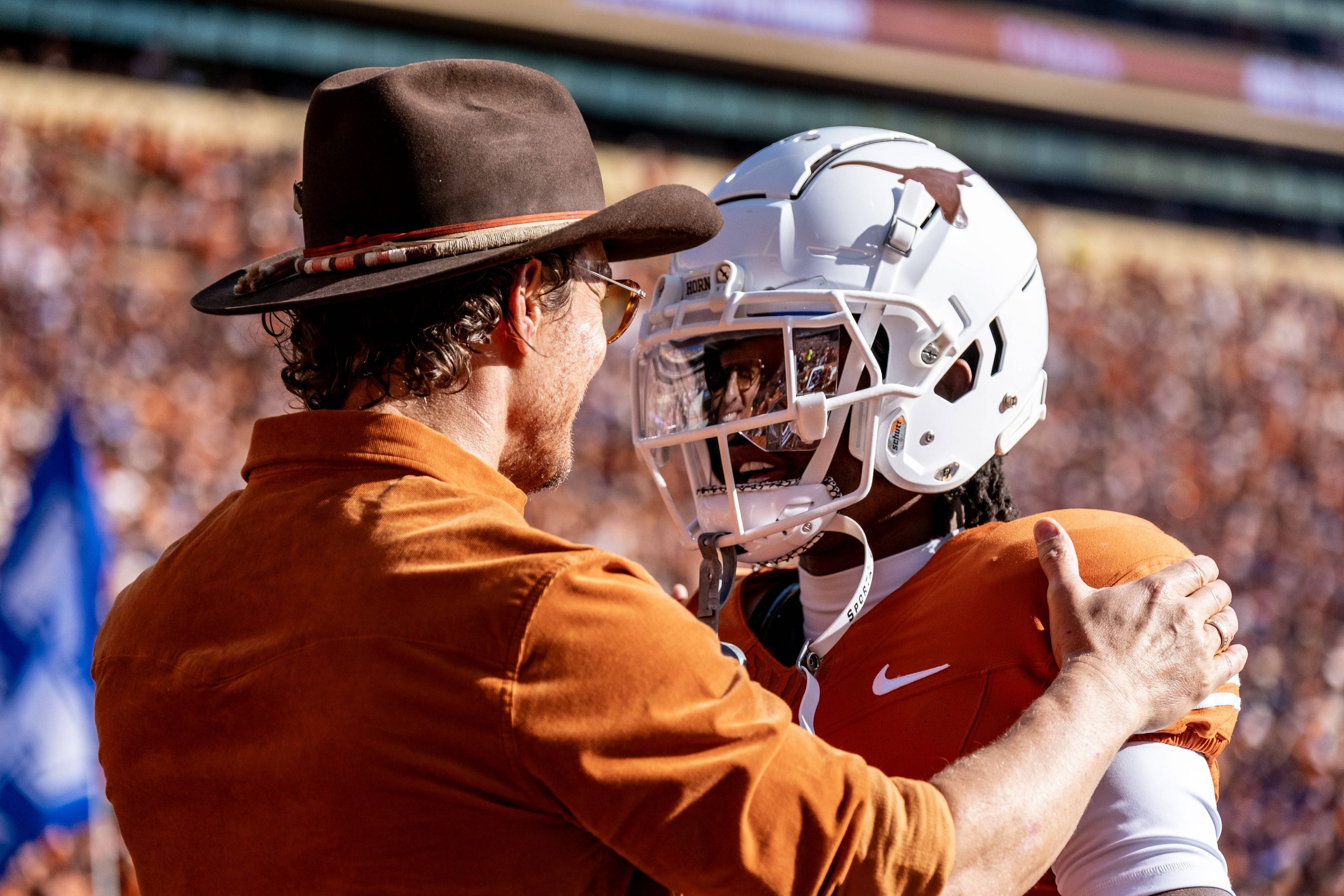 Isaiah Bond - Ein Mann mit braunem Hut und Hemd umarmt Isaiah Bond herzlich. Er trägt eine orange-weiße Uniform und einen Helm mit Longhorn-Logo. Sie stehen auf dem Footballfeld, umgeben von verschwommenen Zuschauern, die die lebhafte Spieltagsatmosphäre einfangen. Diese Beschreibung wurde mit der FootballR KI automatisch generiert.