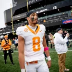 Nico Iamaleava, ein Fußballspieler in einem weiß-orangen Trikot mit der Nummer 8, steht konzentriert auf dem Feld. Er trägt eine rosa Armbinde und sein Gesicht ist bemalt. Hinter ihm fängt ein geschäftiges Stadion voller Zuschauer und Offizieller den Moment trotz des bewölkten Himmels ein. Diese Beschreibung wurde mit der FootballR KI automatisch generiert.