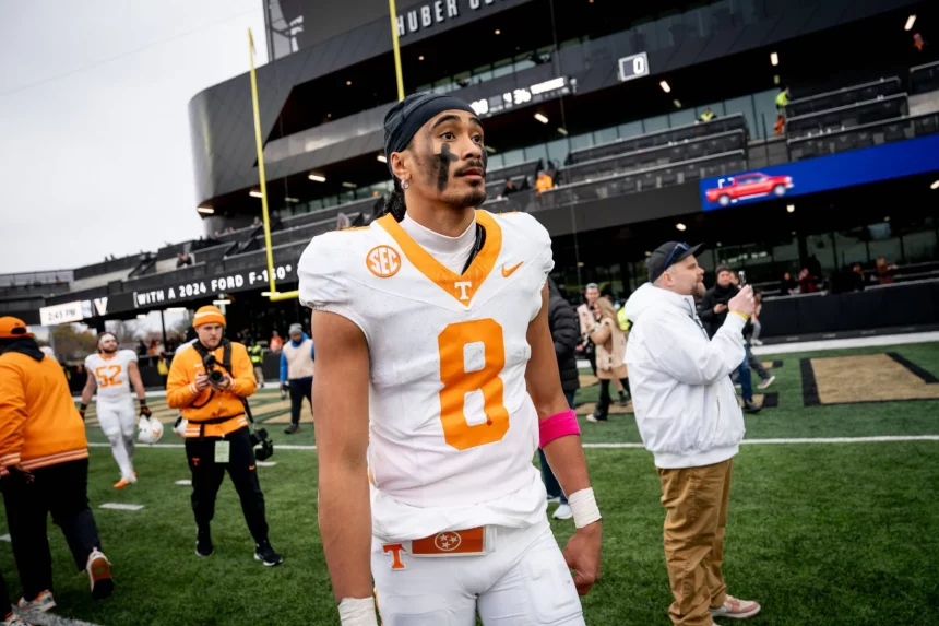 Nico Iamaleava, ein Fußballspieler in einem weiß-orangen Trikot mit der Nummer 8, steht konzentriert auf dem Feld. Er trägt eine rosa Armbinde und sein Gesicht ist bemalt. Hinter ihm fängt ein geschäftiges Stadion voller Zuschauer und Offizieller den Moment trotz des bewölkten Himmels ein. Diese Beschreibung wurde mit der FootballR KI automatisch generiert.