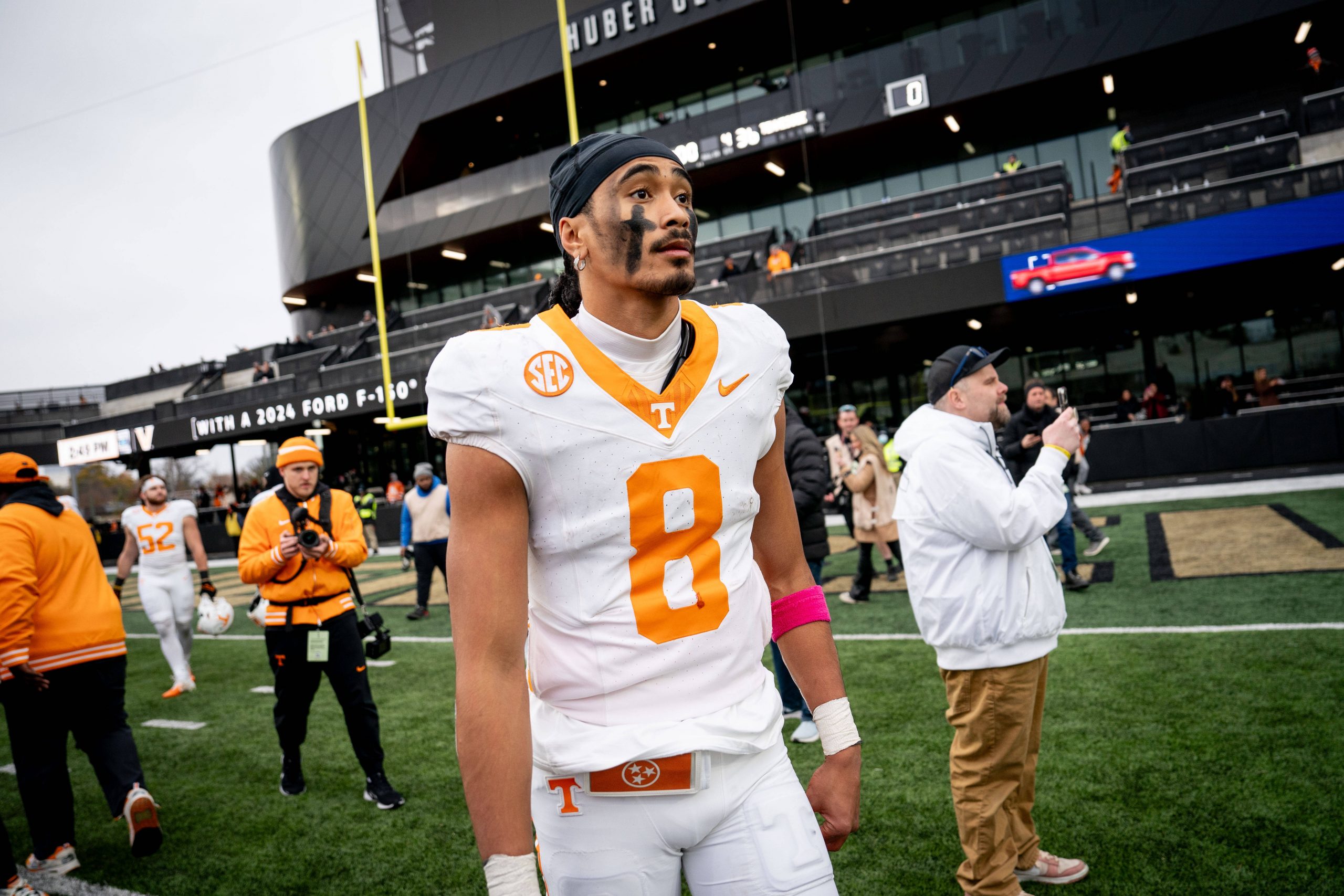 Nico Iamaleava, ein Fußballspieler in einem weiß-orangen Trikot mit der Nummer 8, steht konzentriert auf dem Feld. Er trägt eine rosa Armbinde und sein Gesicht ist bemalt. Hinter ihm fängt ein geschäftiges Stadion voller Zuschauer und Offizieller den Moment trotz des bewölkten Himmels ein. Diese Beschreibung wurde mit der FootballR KI automatisch generiert.