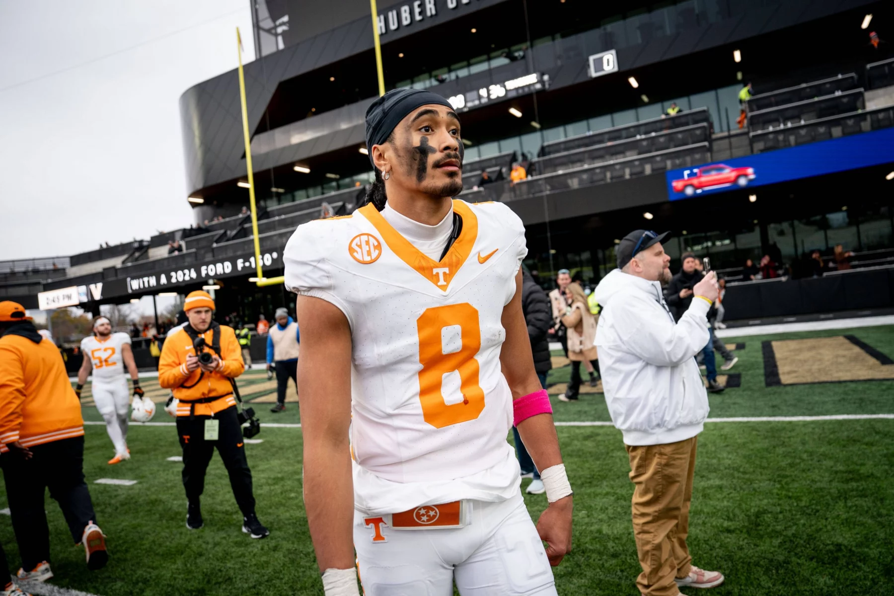 Nico Iamaleava, ein Fußballspieler in einem weiß-orangen Trikot mit der Nummer 8, steht konzentriert auf dem Feld. Er trägt eine rosa Armbinde und sein Gesicht ist bemalt. Hinter ihm fängt ein geschäftiges Stadion voller Zuschauer und Offizieller den Moment trotz des bewölkten Himmels ein. Diese Beschreibung wurde mit der FootballR KI automatisch generiert.