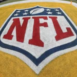 Nahaufnahme des NFL-Logos auf einem Footballfeld. Das schildförmige Logo zeigt einen weißen Football, umgeben von acht Sternen oben, mit „NFL“ in fetten roten Buchstaben auf weißem Hintergrund, alles vor der gelben Endzone – ein Zeichen der Spannung einer bevorstehenden Saison mit 18 Spiele. Diese Beschreibung wurde mit der FootballR KI automatisch generiert.