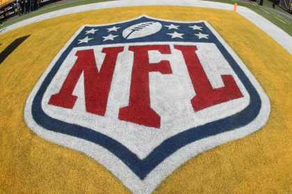 Nahaufnahme des NFL-Logos auf einem Footballfeld. Das schildförmige Logo zeigt einen weißen Football, umgeben von acht Sternen oben, mit „NFL“ in fetten roten Buchstaben auf weißem Hintergrund, alles vor der gelben Endzone – ein Zeichen der Spannung einer bevorstehenden Saison mit 18 Spiele. Diese Beschreibung wurde mit der FootballR KI automatisch generiert.