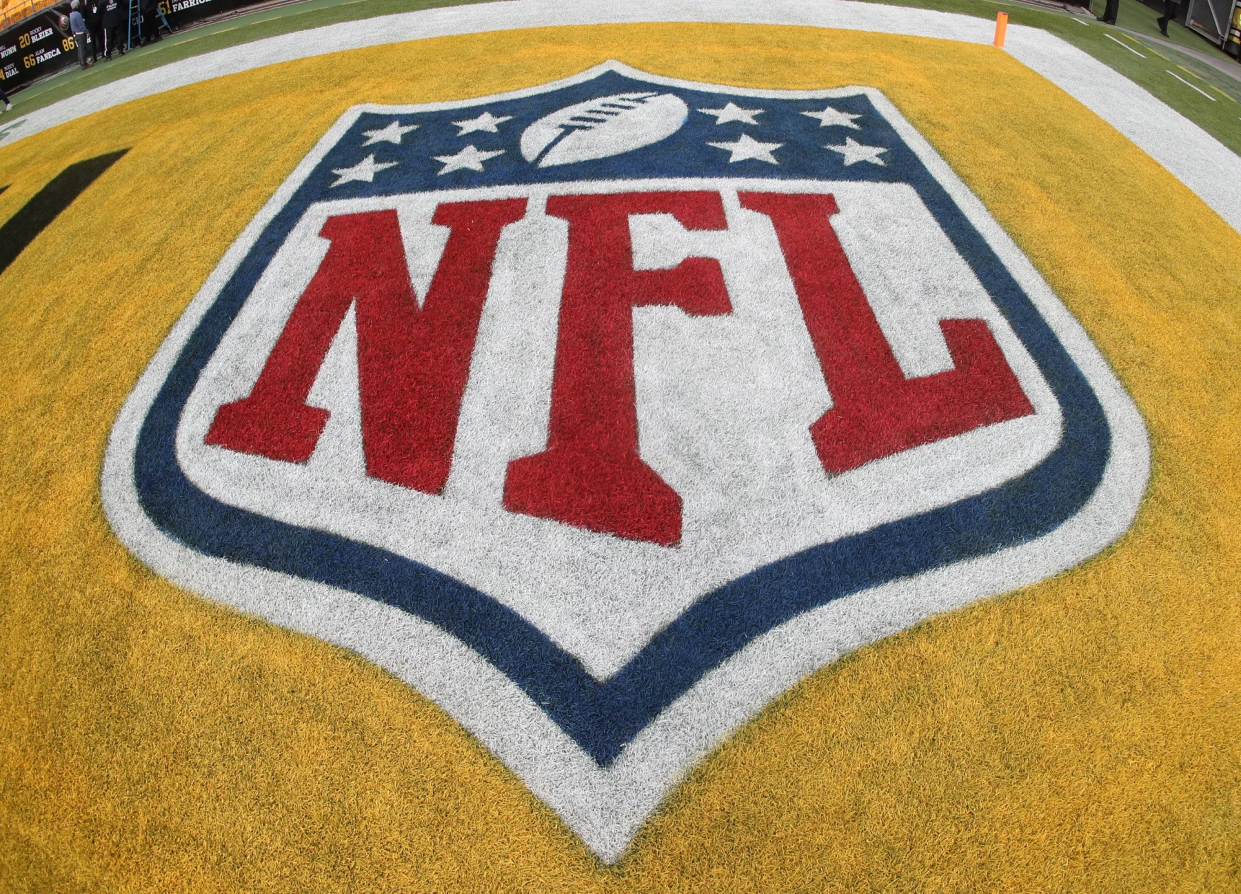 Nahaufnahme des NFL-Logos auf einem Footballfeld. Das schildförmige Logo zeigt einen weißen Football, umgeben von acht Sternen oben, mit „NFL“ in fetten roten Buchstaben auf weißem Hintergrund, alles vor der gelben Endzone – ein Zeichen der Spannung einer bevorstehenden Saison mit 18 Spiele. Diese Beschreibung wurde mit der FootballR KI automatisch generiert.