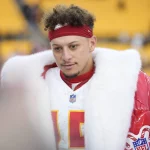 Chiefs vs. Broncos - Kansas City Chiefs Quarterback Patrick Mahomes - Auf dem Feld steht ein Footballspieler mit rotem Stirnband und pelzgefüttertem Mantel. Er trägt ein weißes Trikot mit der Nummer 15. Während er mit einem Reporter, der ein Mikrofon hält, über die letzten Weihnachtsspiele spricht, sind im Hintergrund verschwommene gelbe Stadionsitze zu sehen. Diese Beschreibung wurde mit der FootballR KI automatisch generiert.