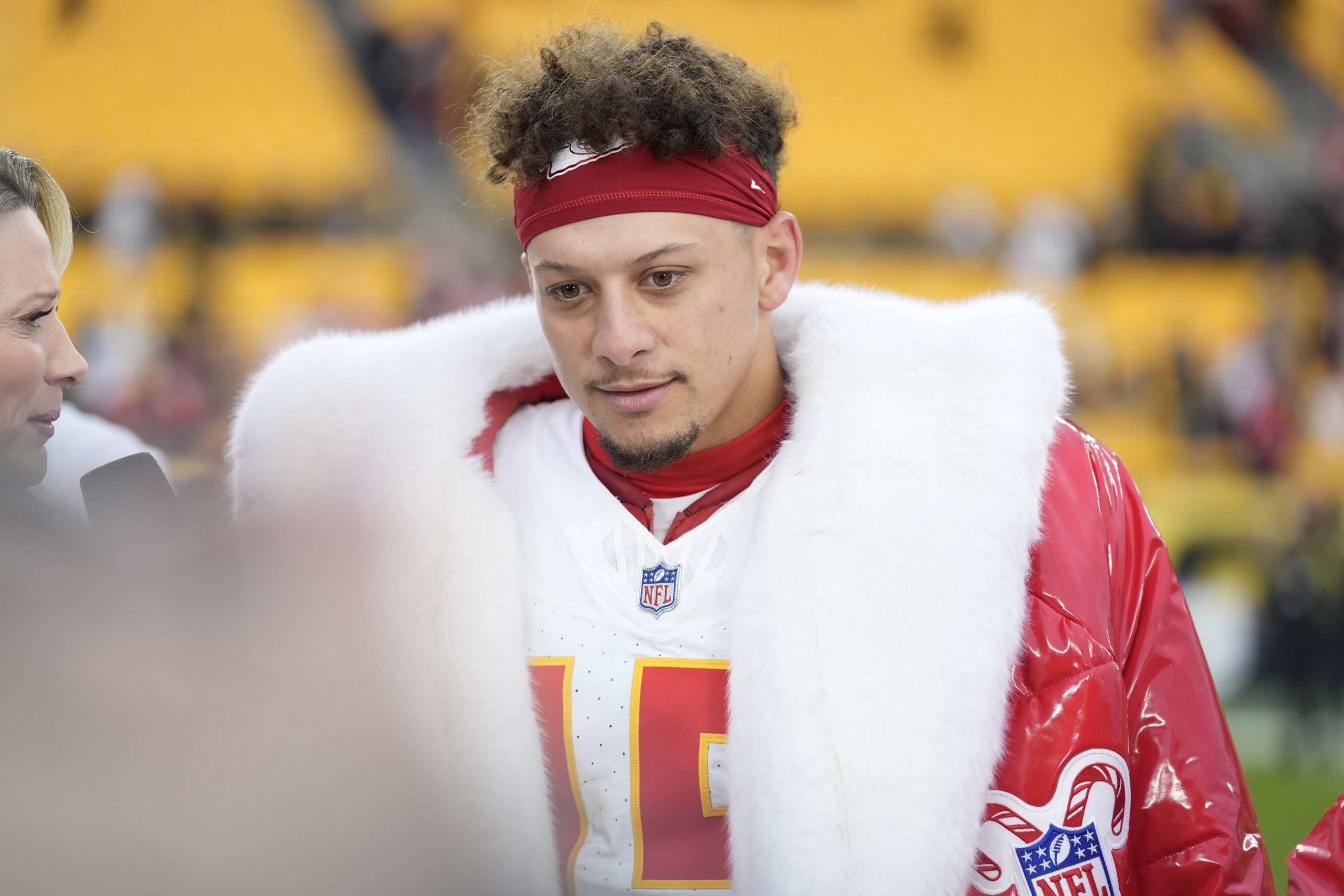 Chiefs vs. Broncos - Kansas City Chiefs Quarterback Patrick Mahomes - Auf dem Feld steht ein Footballspieler mit rotem Stirnband und pelzgefüttertem Mantel. Er trägt ein weißes Trikot mit der Nummer 15. Während er mit einem Reporter, der ein Mikrofon hält, über die letzten Weihnachtsspiele spricht, sind im Hintergrund verschwommene gelbe Stadionsitze zu sehen. Diese Beschreibung wurde mit der FootballR KI automatisch generiert.