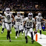 Jack Jones in weißen Trikots jubelt auf einem Spielfeld. Jack Jones läuft mit dem Football in der Hand an der Seitenlinie entlang, seine Teamkollegen rennen ihm hinterher und strecken aufgeregt die Arme in die Höhe. Das Stadion ist voller Zuschauer, im Hintergrund ist ein „Saints“-Schild zu sehen. Diese Beschreibung wurde mit der FootballR KI automatisch generiert.