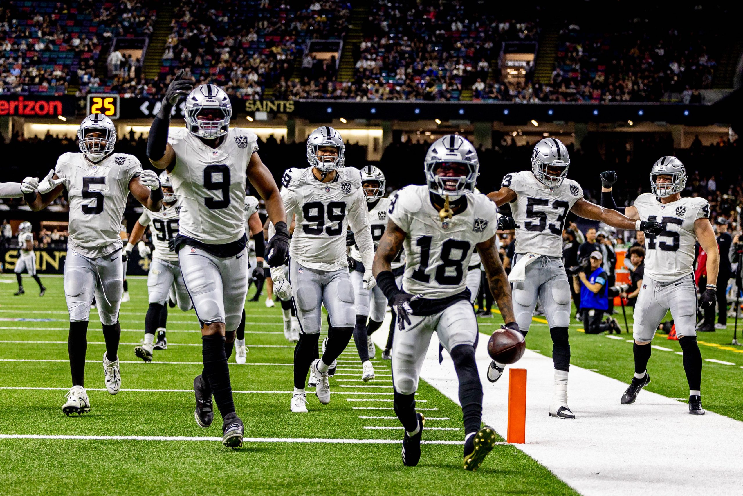 Jack Jones in weißen Trikots jubelt auf einem Spielfeld. Jack Jones läuft mit dem Football in der Hand an der Seitenlinie entlang, seine Teamkollegen rennen ihm hinterher und strecken aufgeregt die Arme in die Höhe. Das Stadion ist voller Zuschauer, im Hintergrund ist ein „Saints“-Schild zu sehen. Diese Beschreibung wurde mit der FootballR KI automatisch generiert.