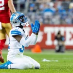 Kerby Joseph, Safety der Detroit Lions, in einer weiß-blauen Uniform, kniet in einer Gebetsgeste auf dem Feld, während ein Spieler der San Francisco 49ers in Rot und Gold während des Spiels weggeht. Diese Beschreibung wurde mit der FootballR KI automatisch generiert.
