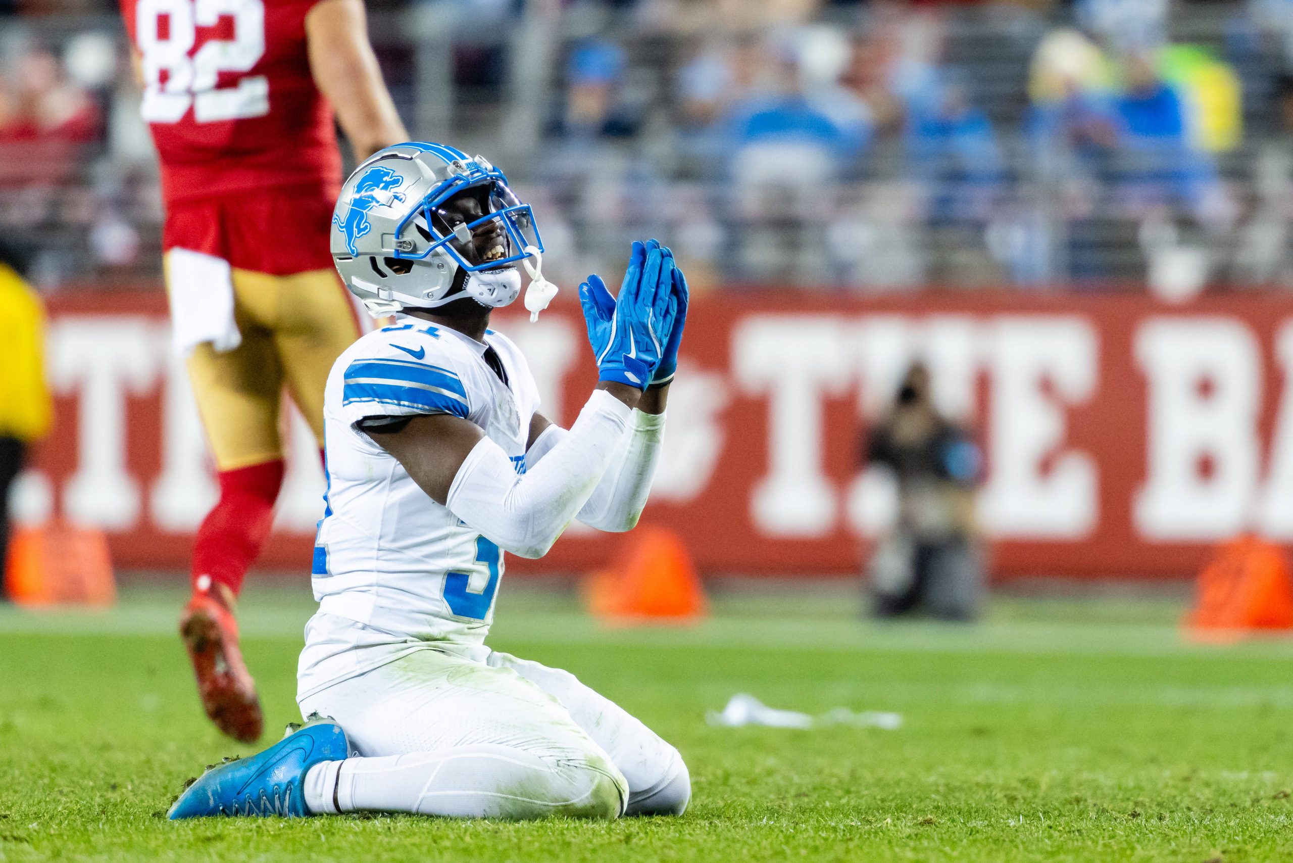 Kerby Joseph, Safety der Detroit Lions, in einer weiß-blauen Uniform, kniet in einer Gebetsgeste auf dem Feld, während ein Spieler der San Francisco 49ers in Rot und Gold während des Spiels weggeht. Diese Beschreibung wurde mit der FootballR KI automatisch generiert.