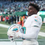 Ein Footballspieler in weißer Miami-Dolphin-Uniform, Tyreek Hill, hält einen passenden Helm mit dem Logo des Teams. Er steht auf dem Footballfeld, im Hintergrund sind Zuschauer zu sehen. Die Szene ist hell, erhellt vom Tageslicht oder von der Stadionbeleuchtung. Diese Beschreibung wurde mit der FootballR KI automatisch generiert.