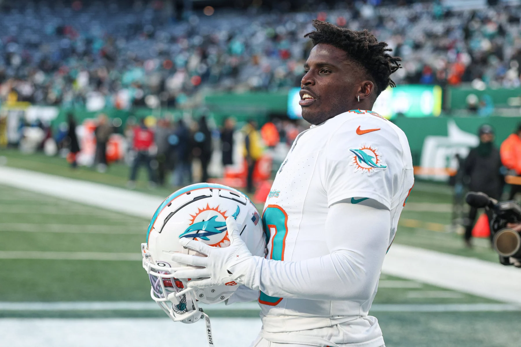 Ein Footballspieler in weißer Miami-Dolphin-Uniform, Tyreek Hill, hält einen passenden Helm mit dem Logo des Teams. Er steht auf dem Footballfeld, im Hintergrund sind Zuschauer zu sehen. Die Szene ist hell, erhellt vom Tageslicht oder von der Stadionbeleuchtung. Diese Beschreibung wurde mit der FootballR KI automatisch generiert.