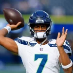 Geno Smith in einem weiß-blauen Trikot mit der Nummer 7, das an einen Klassiker aus Darnold vs. Smith erinnert, bereitet sich auf einen Footballwurf vor. Mit blauem Helm und Visier steht er hochkonzentriert vor dem Hintergrund eines verschwommenen Stadions, in dem das Spielgeschehen pulsiert. Diese Beschreibung wurde mit der FootballR KI automatisch generiert.