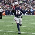 Joe Milton III in Marineblau und Rot mit der Nummer 19, der Joe Milton ähnelt, sprintet über das Feld. Mit Helm hält er den Ball in der rechten Hand. Die Zuschauer beobachten gespannt von der Tribüne aus, wie sich ein Schiedsrichter im gestreiften Trikot links positioniert. Diese Beschreibung wurde mit der FootballR KI automatisch generiert.