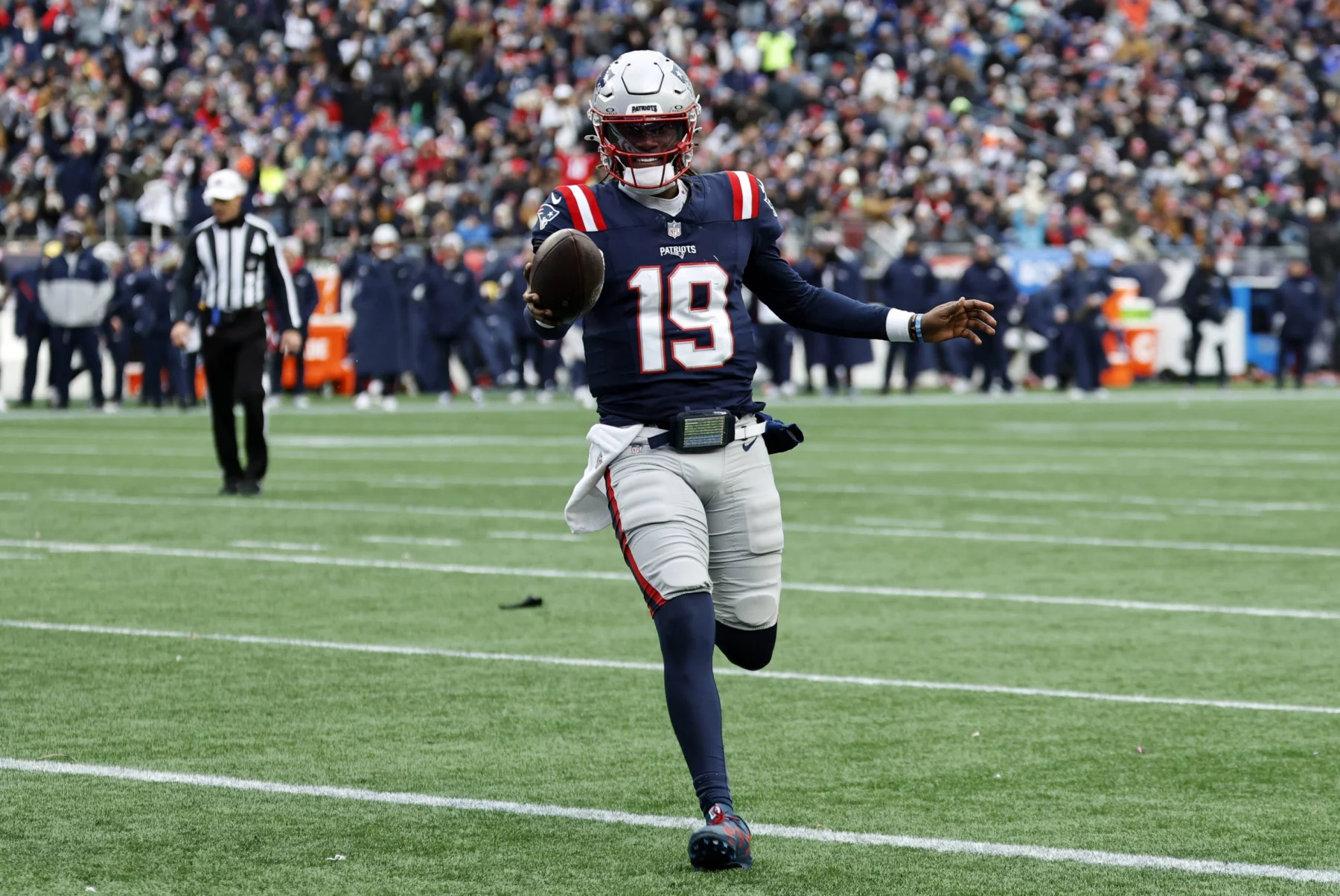 Joe Milton III in Marineblau und Rot mit der Nummer 19, der Joe Milton ähnelt, sprintet über das Feld. Mit Helm hält er den Ball in der rechten Hand. Die Zuschauer beobachten gespannt von der Tribüne aus, wie sich ein Schiedsrichter im gestreiften Trikot links positioniert. Diese Beschreibung wurde mit der FootballR KI automatisch generiert.