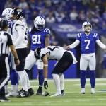 Ein Footballspiel läuft. Spieler tragen blau-weiße Trikots. Kicker Matt Gay mit der Nummer 7 steht gelassen da. Die gegnerische Mannschaft, schwarz-weiß gekleidet, hat die Nummer 44 nach vorne gebeugt. Links blickt der Schiedsrichter in schwarz-weißen Streifen vor einer verschwommenen Zuschauerkulisse auf das Geschehen. Diese Beschreibung wurde mit der FootballR KI automatisch generiert.