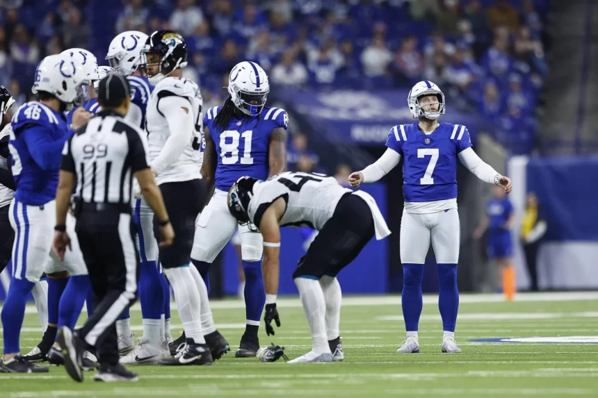 Ein Footballspiel läuft. Spieler tragen blau-weiße Trikots. Kicker Matt Gay mit der Nummer 7 steht gelassen da. Die gegnerische Mannschaft, schwarz-weiß gekleidet, hat die Nummer 44 nach vorne gebeugt. Links blickt der Schiedsrichter in schwarz-weißen Streifen vor einer verschwommenen Zuschauerkulisse auf das Geschehen. Diese Beschreibung wurde mit der FootballR KI automatisch generiert.