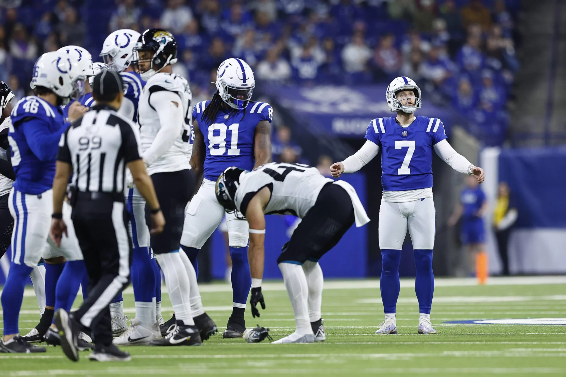 Ein Footballspiel läuft. Spieler tragen blau-weiße Trikots. Kicker Matt Gay mit der Nummer 7 steht gelassen da. Die gegnerische Mannschaft, schwarz-weiß gekleidet, hat die Nummer 44 nach vorne gebeugt. Links blickt der Schiedsrichter in schwarz-weißen Streifen vor einer verschwommenen Zuschauerkulisse auf das Geschehen. Diese Beschreibung wurde mit der FootballR KI automatisch generiert.