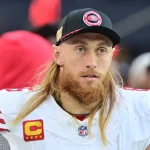 Fantasy Football Woche 7 - Ein Footballspieler mit langen blonden Haaren und rötlichem Bart trägt die Uniform der San Francisco 49ers und eine schwarze Kappe. Auf seinem weißen Trikot trägt er – wie Star-Tight-End George Kittle – einen Kapitänsaufnäher und blickt mit neutralem Gesichtsausdruck nach vorn. Diese Beschreibung wurde mit der FootballR KI automatisch generiert.