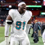 Der Footballspieler der Miami Dolphins, Emmanuel Ogbah, trägt die Trikotnummer 91 und lächelt breit auf dem Spielfeld. Er trägt eine komplett weiße Uniform mit blaugrünen Akzenten und ein weißes Stirnband. Im Hintergrund sind Stadionsitze und andere Spieler zu sehen. Diese Beschreibung wurde mit der FootballR KI automatisch generiert.