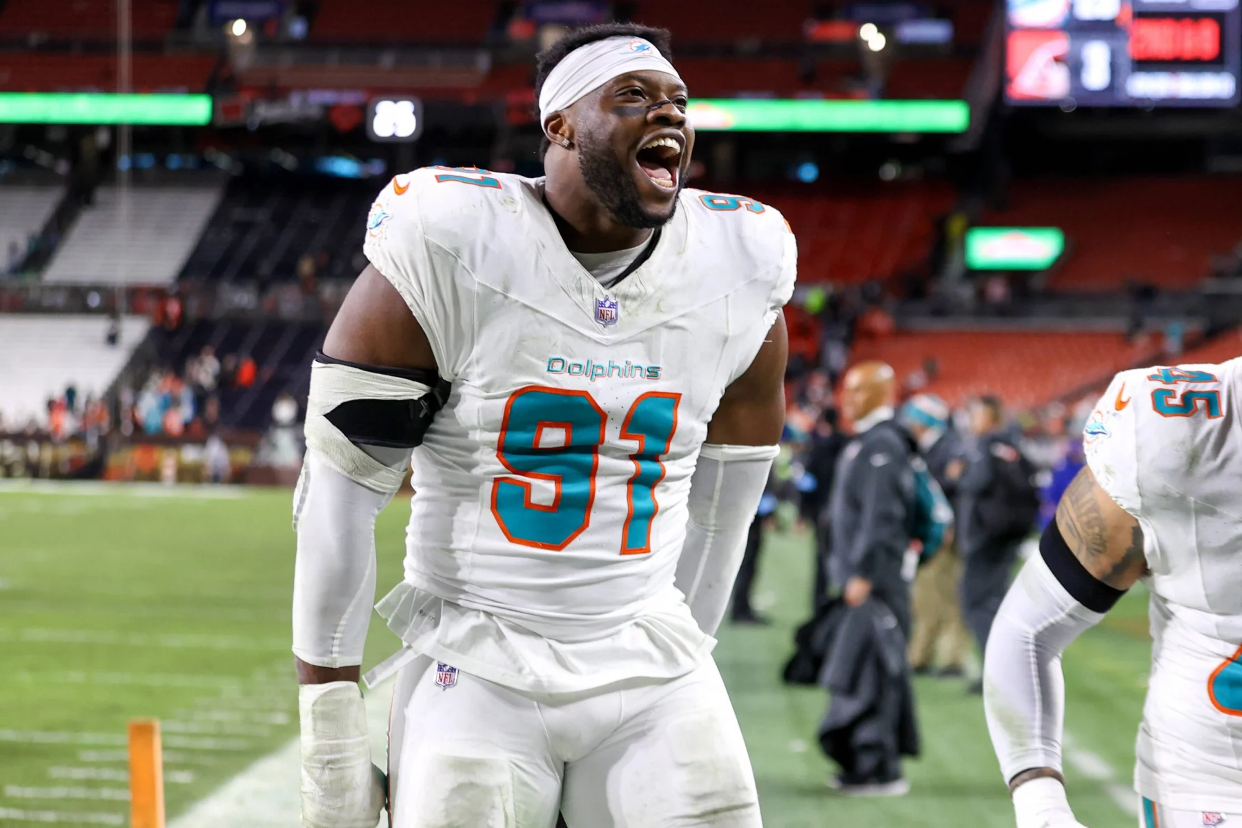 Der Footballspieler der Miami Dolphins, Emmanuel Ogbah, trägt die Trikotnummer 91 und lächelt breit auf dem Spielfeld. Er trägt eine komplett weiße Uniform mit blaugrünen Akzenten und ein weißes Stirnband. Im Hintergrund sind Stadionsitze und andere Spieler zu sehen. Diese Beschreibung wurde mit der FootballR KI automatisch generiert.