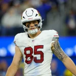 Footballspieler Trey McBride steht selbstbewusst auf dem Spielfeld. Er trägt ein weißes Trikot mit der Nummer 85 und einen Helm mit Vogellogo. Schwarze Augenfarbe unterstreicht seine Konzentration, während ein auffälliges Tattoo seinen rechten Arm ziert. Hinter ihm sorgen die verschwommene Menge und der leuchtend blaue LED-Bildschirm für eine elektrisierende Atmosphäre. Diese Beschreibung wurde mit der FootballR KI automatisch generiert.