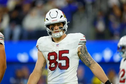 Footballspieler Trey McBride steht selbstbewusst auf dem Spielfeld. Er trägt ein weißes Trikot mit der Nummer 85 und einen Helm mit Vogellogo. Schwarze Augenfarbe unterstreicht seine Konzentration, während ein auffälliges Tattoo seinen rechten Arm ziert. Hinter ihm sorgen die verschwommene Menge und der leuchtend blaue LED-Bildschirm für eine elektrisierende Atmosphäre. Diese Beschreibung wurde mit der FootballR KI automatisch generiert.