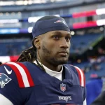 Joe Milton III in der Uniform der New England Patriots steht auf einem Spielfeld. Er ähnelt Joe Milton mit schulterlangem Haar unter einem blauen Stirnband. Er trägt ein blaues Trikot mit rot-weißen Ärmeln vor einer Stadionkulisse mit verschwommenen Zuschauern und Werbebannern. Diese Beschreibung wurde mit der FootballR KI automatisch generiert.