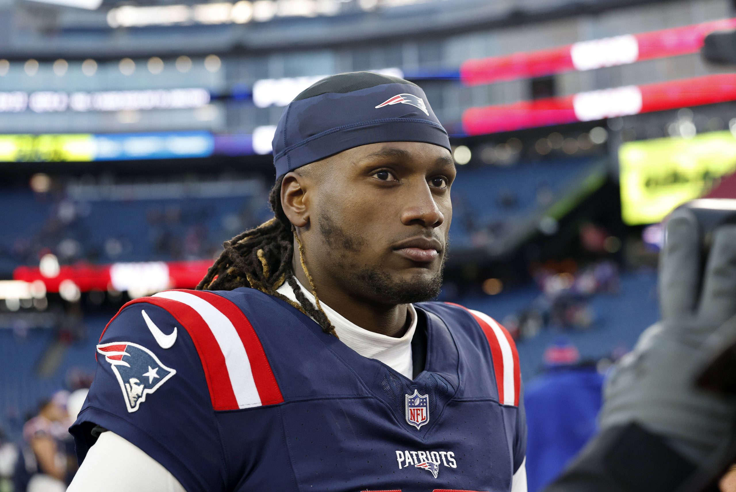 Joe Milton III in der Uniform der New England Patriots steht auf einem Spielfeld. Er ähnelt Joe Milton mit schulterlangem Haar unter einem blauen Stirnband. Er trägt ein blaues Trikot mit rot-weißen Ärmeln vor einer Stadionkulisse mit verschwommenen Zuschauern und Werbebannern. Diese Beschreibung wurde mit der FootballR KI automatisch generiert.