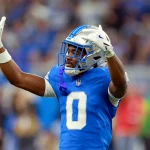 Detroit-Lion-Spieler Terrion Arnold in blauer Uniform und silbernem Helm streckt während eines Spiels beide Arme mit gebeugten Händen in die Höhe. Mit der Nummer 0 sticht er aus der verschwommenen Menge hervor und fängt die aufregende Stadionatmosphäre ein. Diese Beschreibung wurde mit der FootballR KI automatisch generiert.