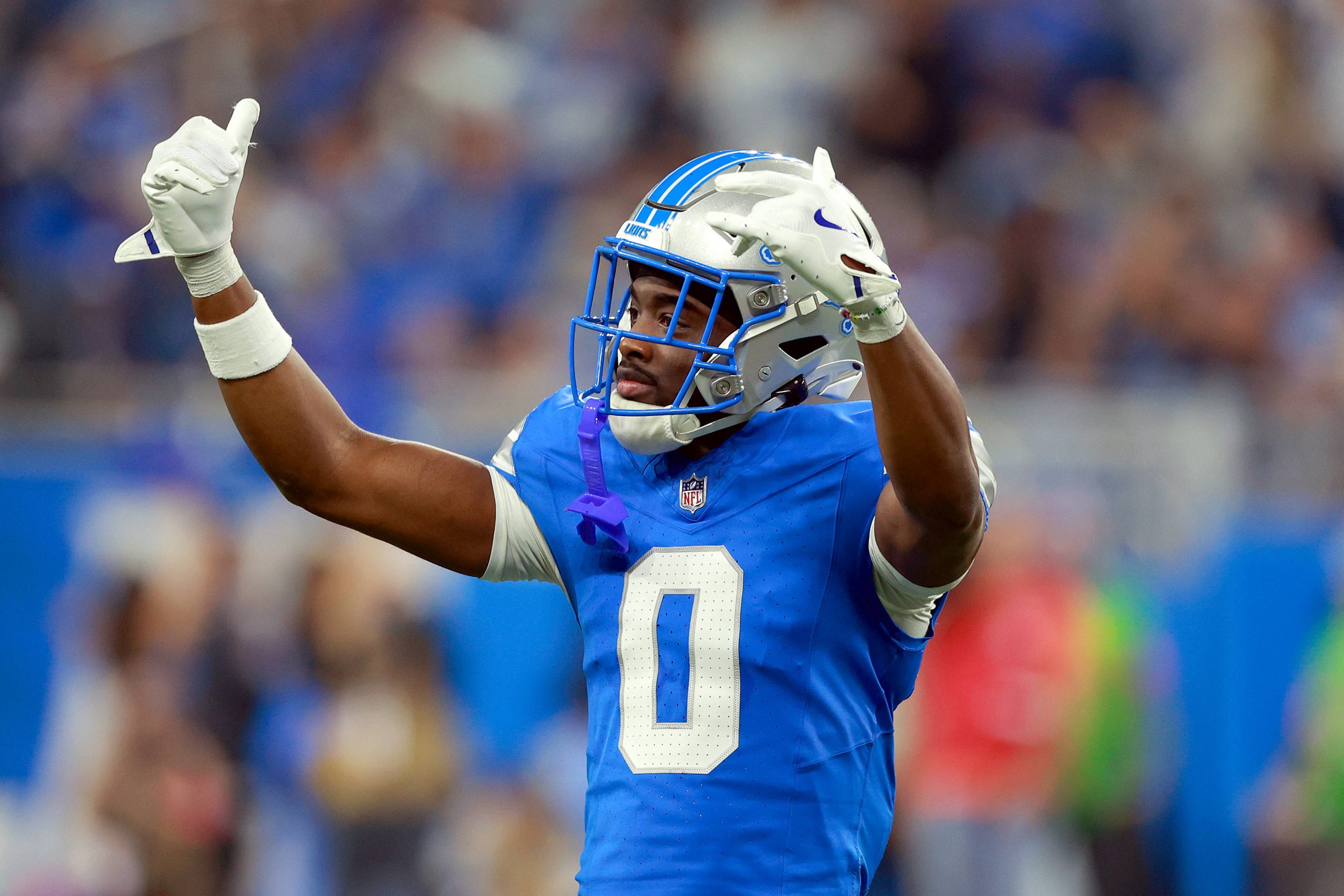 Detroit-Lion-Spieler Terrion Arnold in blauer Uniform und silbernem Helm streckt während eines Spiels beide Arme mit gebeugten Händen in die Höhe. Mit der Nummer 0 sticht er aus der verschwommenen Menge hervor und fängt die aufregende Stadionatmosphäre ein. Diese Beschreibung wurde mit der FootballR KI automatisch generiert.