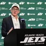 Woody Johnson in Anzug und Jets-Kappe spricht auf einem Podium mit der Aufschrift „Atlantic Health New York Jets Training Center“. Der Hintergrund ist grün, mit mehreren „Jets“-Logos und Sponsorennamen, darunter „Cash App“ und „Atlantic Health System“. Ein Mikrofon mit blauer Abdeckung ist zu sehen. Diese Beschreibung wurde mit der FootballR KI automatisch generiert.