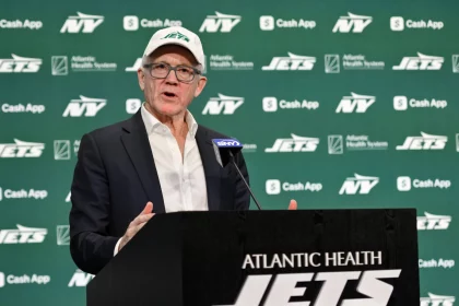 Woody Johnson in Anzug und Jets-Kappe spricht auf einem Podium mit der Aufschrift „Atlantic Health New York Jets Training Center“. Der Hintergrund ist grün, mit mehreren „Jets“-Logos und Sponsorennamen, darunter „Cash App“ und „Atlantic Health System“. Ein Mikrofon mit blauer Abdeckung ist zu sehen. Diese Beschreibung wurde mit der FootballR KI automatisch generiert.