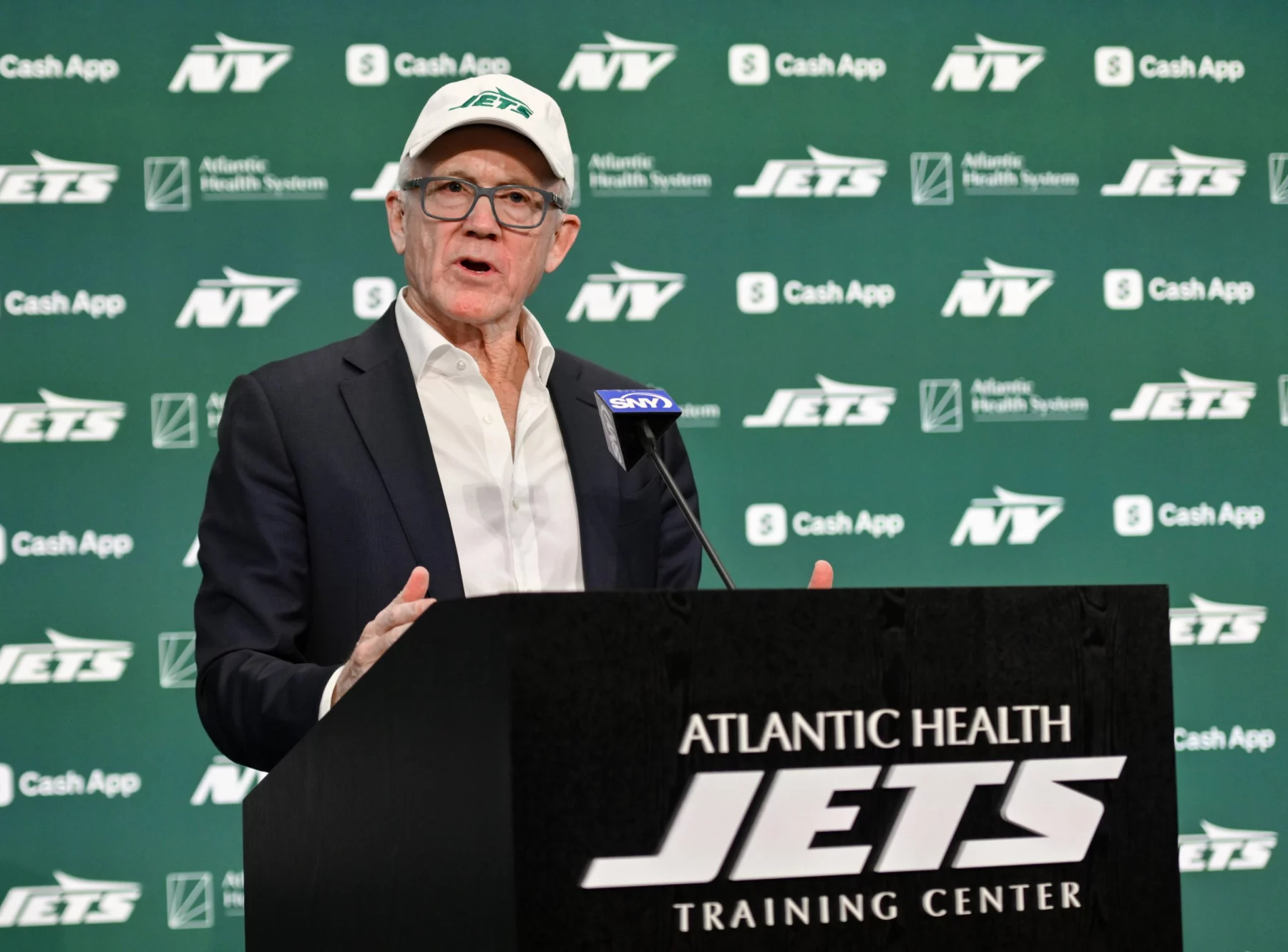 Woody Johnson in Anzug und Jets-Kappe spricht auf einem Podium mit der Aufschrift „Atlantic Health New York Jets Training Center“. Der Hintergrund ist grün, mit mehreren „Jets“-Logos und Sponsorennamen, darunter „Cash App“ und „Atlantic Health System“. Ein Mikrofon mit blauer Abdeckung ist zu sehen. Diese Beschreibung wurde mit der FootballR KI automatisch generiert.