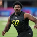 Darius Alexander, ein Footballspieler, rennt konzentriert und entschlossen über das Feld. In einem schwarzen ärmellosen Trikot mit der Nummer „02“ und „DL“ in Gelb bewegt er sich vor der verschwommenen Kulisse eines lebendigen Stadions voller bunter Banner über einem makellosen Rasenfeld. Diese Beschreibung wurde mit der FootballR KI automatisch generiert.