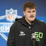 Grey Zabel, ein Mann mit kurzen braunen Haaren und Schnurrbart, steht am Mikrofon und trägt einen schwarzen New Era Hoodie mit neongrünem „50 OL“-Schriftzug. Hinter ihm ist ein blauer NFL Combine-Hintergrund zu sehen, auf dem das NFL-Logo deutlich zu sehen ist. Diese Beschreibung wurde mit der FootballR KI automatisch generiert.