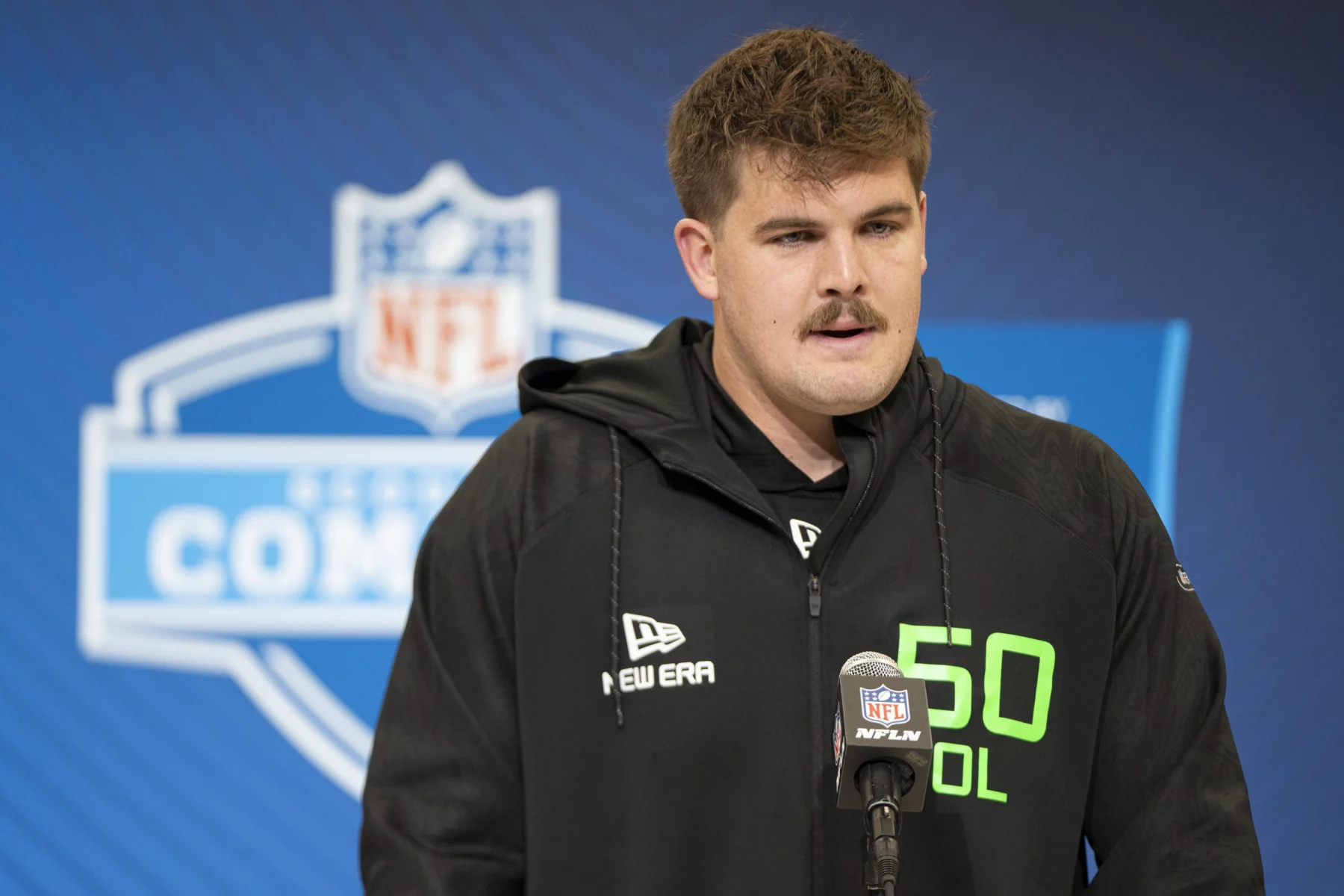 Grey Zabel, ein Mann mit kurzen braunen Haaren und Schnurrbart, steht am Mikrofon und trägt einen schwarzen New Era Hoodie mit neongrünem „50 OL“-Schriftzug. Hinter ihm ist ein blauer NFL Combine-Hintergrund zu sehen, auf dem das NFL-Logo deutlich zu sehen ist. Diese Beschreibung wurde mit der FootballR KI automatisch generiert.