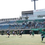 In einem Freiluftstadion findet ein Flag Football Spiel statt, bei dem Spieler in schwarz-weißen Trikots gegen Spieler in grünen Trikots antreten. Vereinzelte Zuschauer besetzen die Tribünen unter klarem Himmel, während eine Anzeigetafel mit Werbung darüber thront und die Atmosphäre dieses spannenden Flag-Football-Spiels einfängt. Diese Beschreibung wurde mit der FootballR KI automatisch generiert.