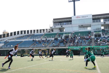 In einem Freiluftstadion findet ein Flag Football Spiel statt, bei dem Spieler in schwarz-weißen Trikots gegen Spieler in grünen Trikots antreten. Vereinzelte Zuschauer besetzen die Tribünen unter klarem Himmel, während eine Anzeigetafel mit Werbung darüber thront und die Atmosphäre dieses spannenden Flag-Football-Spiels einfängt. Diese Beschreibung wurde mit der FootballR KI automatisch generiert.