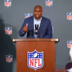 NFL gegen unsportliches Verhalten: Troy Vincent im blauen Anzug spricht an einem mit NFL-Logos geschmückten Podium und verkörpert Sportsgeist. Er gestikuliert selbstbewusst mit der rechten Hand und hält mit der linken ein Mikrofon. Im Hintergrund wiederholen sich NFL-Logos. Aufmerksame Zuhörer im Vordergrund reflektieren sein konzentriertes Auftreten. Diese Beschreibung wurde mit der FootballR KI automatisch generiert.