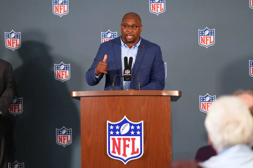 NFL gegen unsportliches Verhalten: Troy Vincent im blauen Anzug spricht an einem mit NFL-Logos geschmückten Podium und verkörpert Sportsgeist. Er gestikuliert selbstbewusst mit der rechten Hand und hält mit der linken ein Mikrofon. Im Hintergrund wiederholen sich NFL-Logos. Aufmerksame Zuhörer im Vordergrund reflektieren sein konzentriertes Auftreten. Diese Beschreibung wurde mit der FootballR KI automatisch generiert.