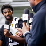 Shedeur Sanders im schwarzen Hemd und mit einem Handtuch über den Schultern unterhält sich mit zwei anderen, die ESPN-Mikrofone halten. Vor dem Hintergrund einer scheinbar öffentlichen Titans-Veranstaltung verschwimmen die Details. Diese Beschreibung wurde mit der FootballR KI automatisch generiert.