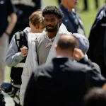 Shedeur Sanders in einer großen weißen Jacke und einer dicken Diamantenkette steht im Freien inmitten einer Gruppe von Menschen beim NFL Draft 2025. Er hat kurzes, lockiges Haar und einen gestutzten Bart. Im unscharfen Hintergrund ist die Kameraausrüstung zu sehen. Diese Beschreibung wurde mit der FootballR KI automatisch generiert.