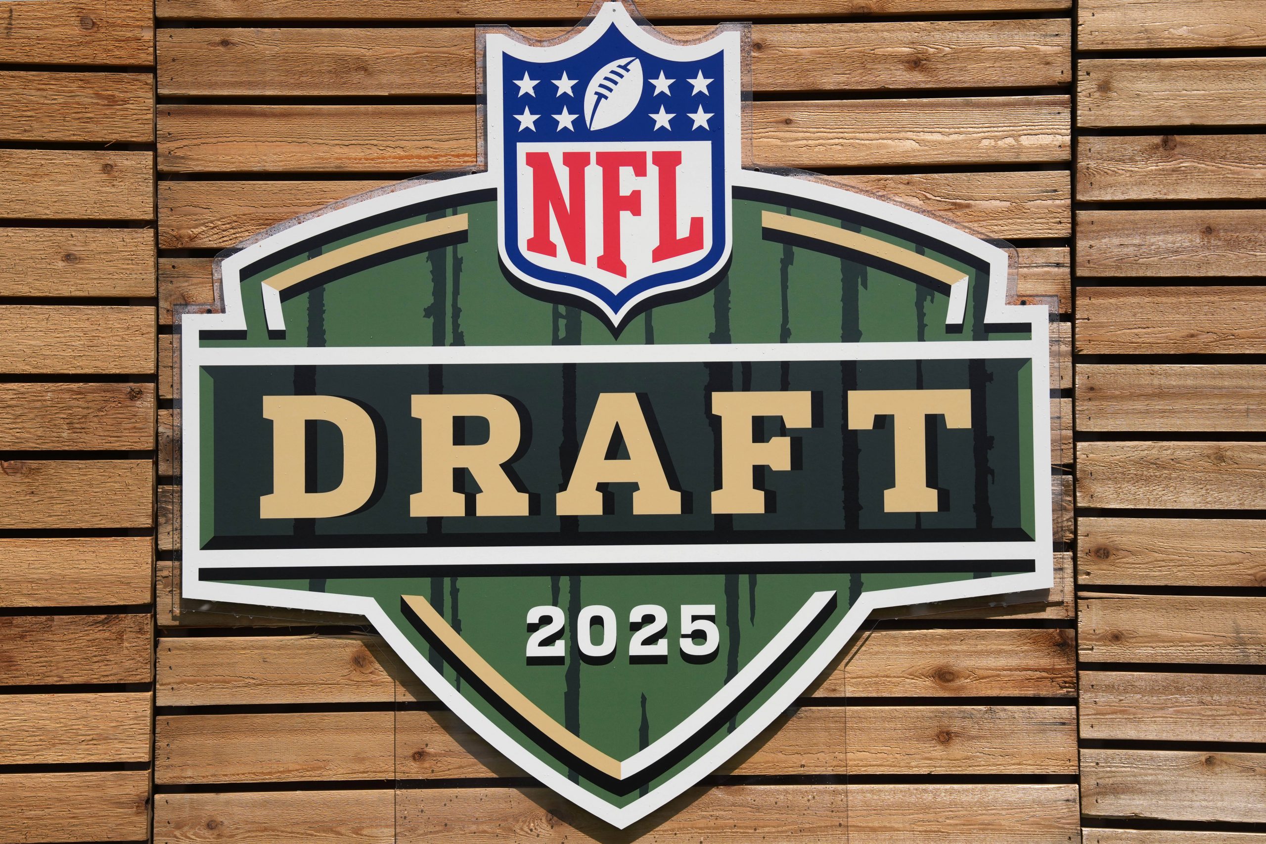 NFL Draft - Ein großes, schildförmiges Schild mit dem NFL-Logo darüber und „DRAFT“ in fetten Buchstaben sowie „2025“ darunter steht in Grün, Gold und Weiß an einer Holzplankenwand und unterstreicht die Spannung der Verpflichtung von nicht gedrafteten Free Agents. Diese Beschreibung wurde mit der FootballR KI automatisch generiert.