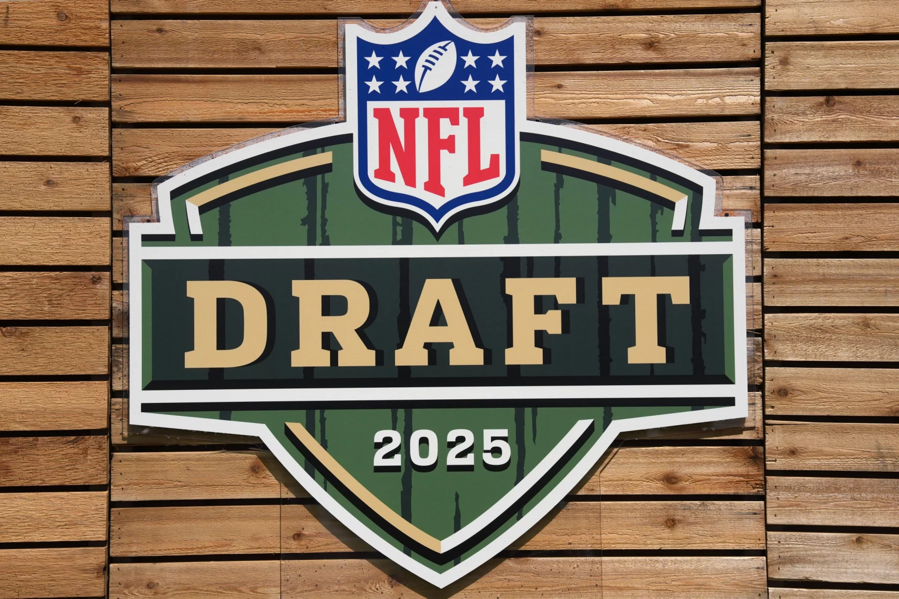 NFL Draft - Ein großes, schildförmiges Schild mit dem NFL-Logo darüber und „DRAFT“ in fetten Buchstaben sowie „2025“ darunter steht in Grün, Gold und Weiß an einer Holzplankenwand und unterstreicht die Spannung der Verpflichtung von nicht gedrafteten Free Agents. Diese Beschreibung wurde mit der FootballR KI automatisch generiert.