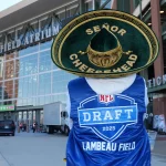 Eine Person in einem blauen Lambeau Field-Umhang (NFL Draft 2025) und einem weißen Kapuzenpullover steht vor dem Lambeau Field. Sie trägt eine große grüne „Señor Cheesehead“-Mütze mit einer goldenen Käsescheibe und posiert in der Nähe des Stadioneingangs und der Ticketschalter. Diese Beschreibung wurde mit der FootballR KI automatisch generiert.