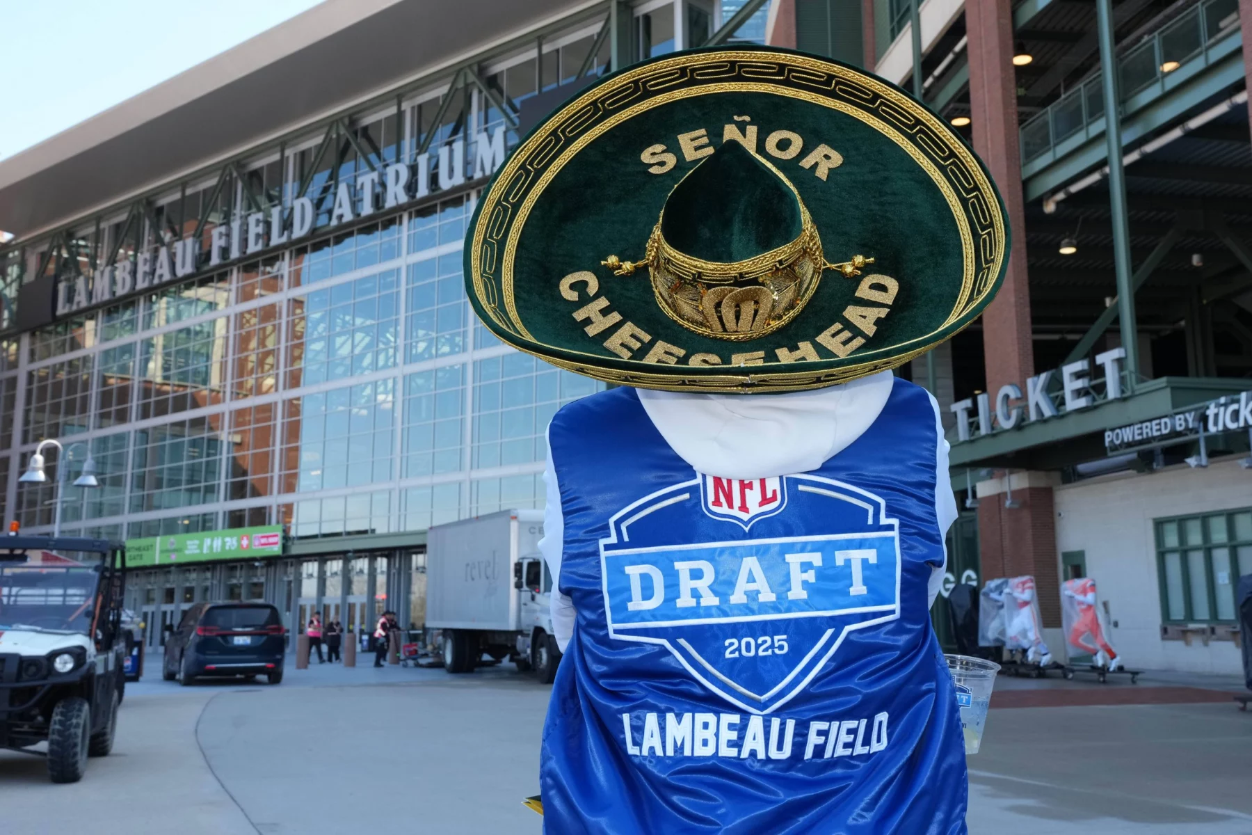 Eine Person in einem blauen Lambeau Field-Umhang (NFL Draft 2025) und einem weißen Kapuzenpullover steht vor dem Lambeau Field. Sie trägt eine große grüne „Señor Cheesehead“-Mütze mit einer goldenen Käsescheibe und posiert in der Nähe des Stadioneingangs und der Ticketschalter. Diese Beschreibung wurde mit der FootballR KI automatisch generiert.