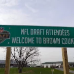 NFL Draft 2025 - Cleveland Browns und New York Giants planen Trades. Auf einem grünen Schild am Straßenrand mit dem Logo des NFL Draft 2025 steht „Willkommen in Brown County, Teilnehmer des NFL Draft“. Das Schild ist auf Holzpfosten montiert und steht vor einer Kulisse aus Gebäuden, Gras und einem kahlen Baum. Diese Beschreibung wurde mit der FootballR KI automatisch generiert.