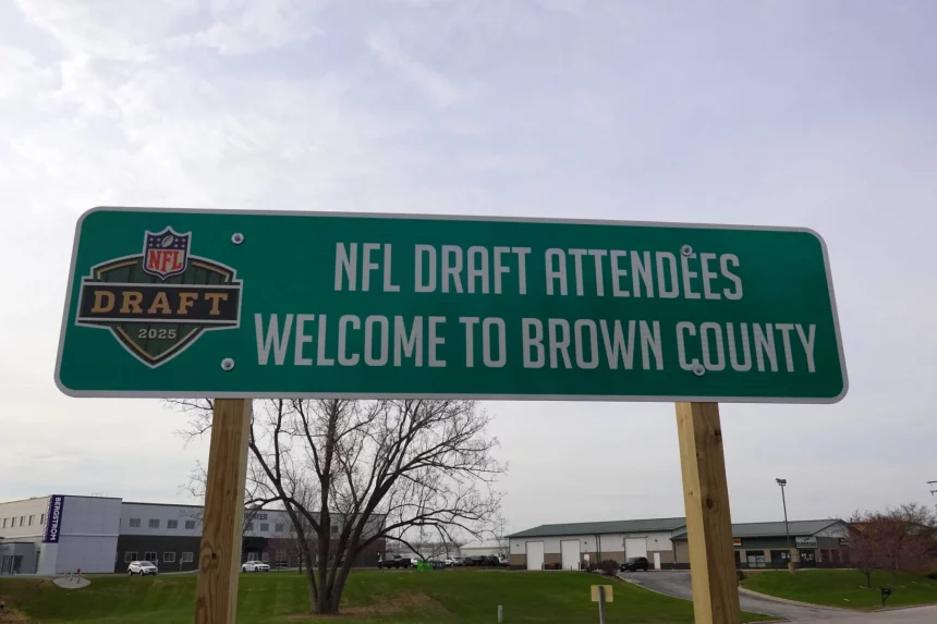 NFL Draft 2025 - Cleveland Browns und New York Giants planen Trades. Auf einem grünen Schild am Straßenrand mit dem Logo des NFL Draft 2025 steht „Willkommen in Brown County, Teilnehmer des NFL Draft“. Das Schild ist auf Holzpfosten montiert und steht vor einer Kulisse aus Gebäuden, Gras und einem kahlen Baum. Diese Beschreibung wurde mit der FootballR KI automatisch generiert.